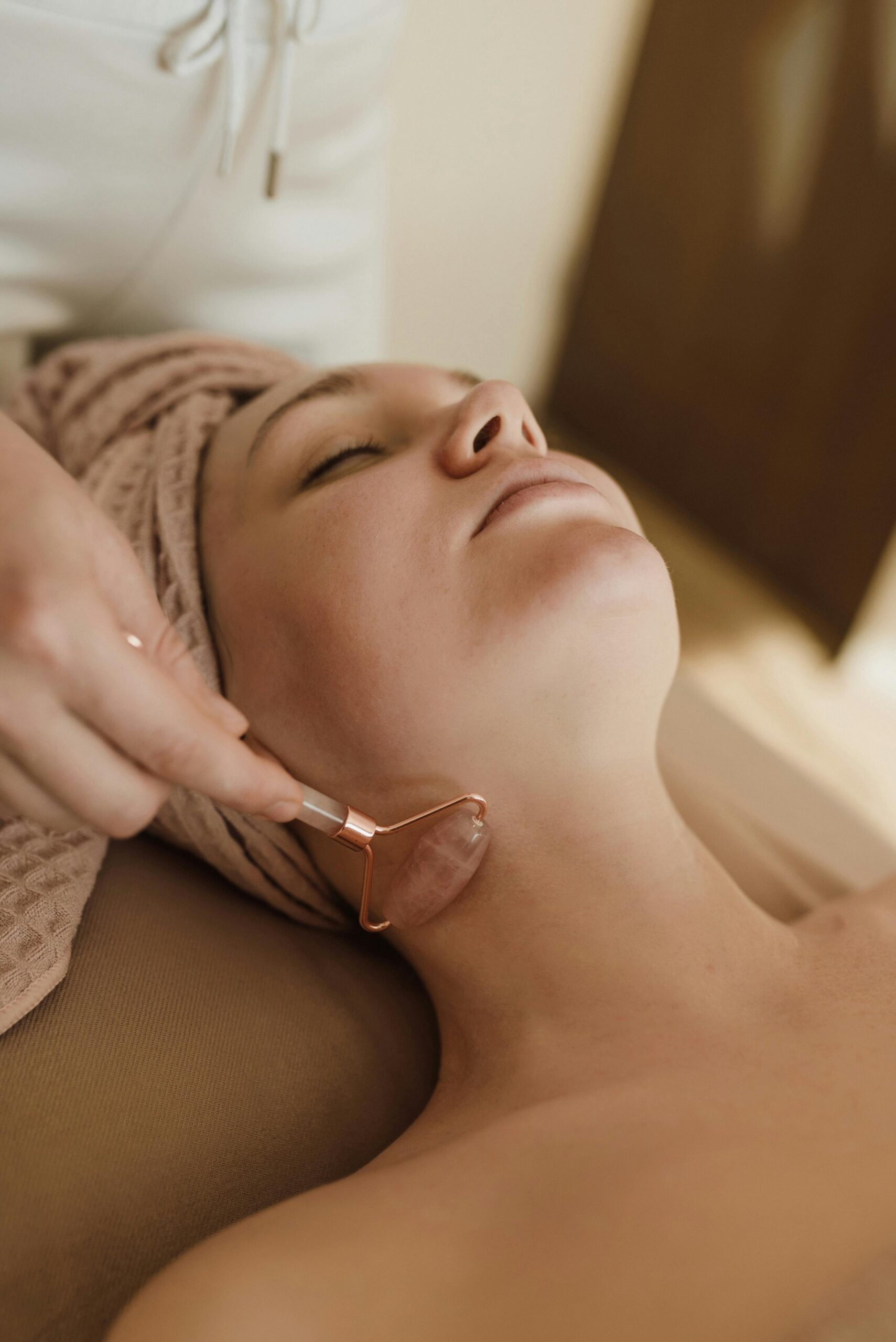 Esthetician glides a rose‑quartz roller along a woman’s neck and jawline during facial treatment.