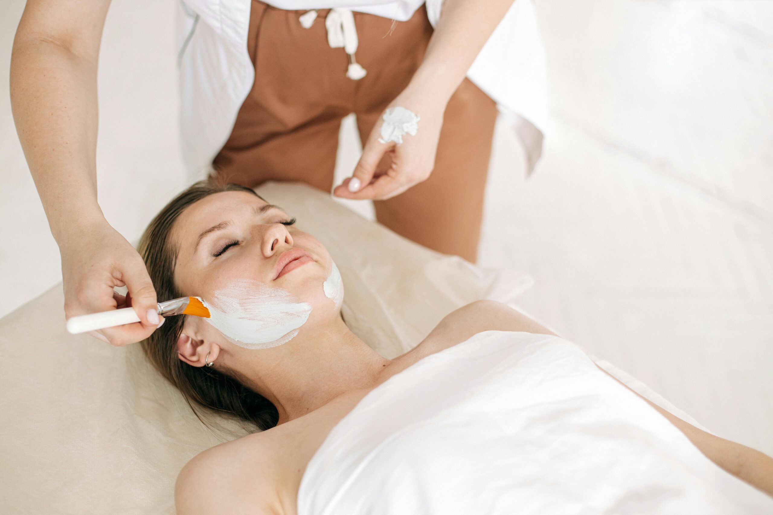 Facialist spreads thick white mask on reclining woman’s cheeks with a flat brush.