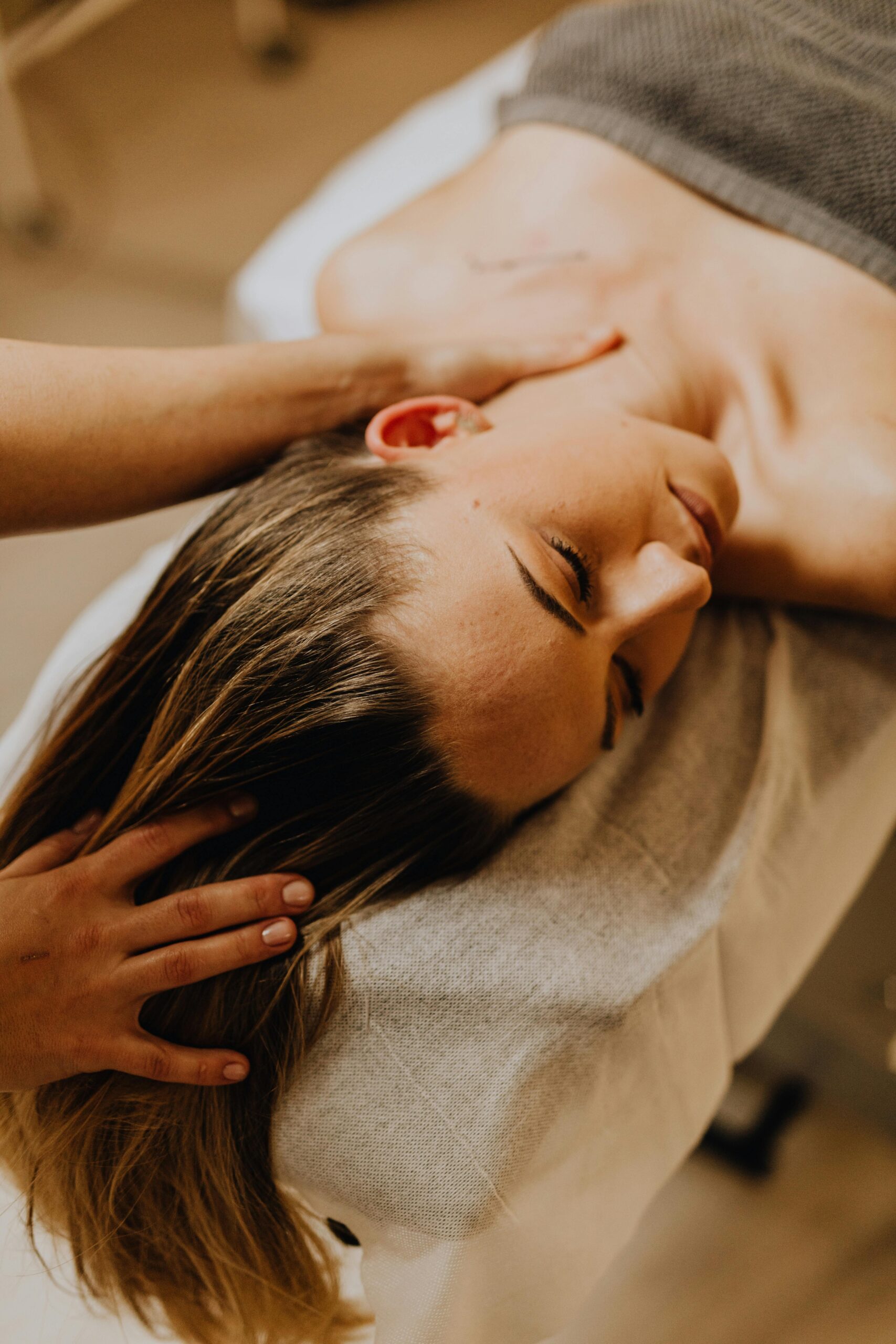 Woman lies side‑head on a cushioned table while a therapist massages her neck and scalp.