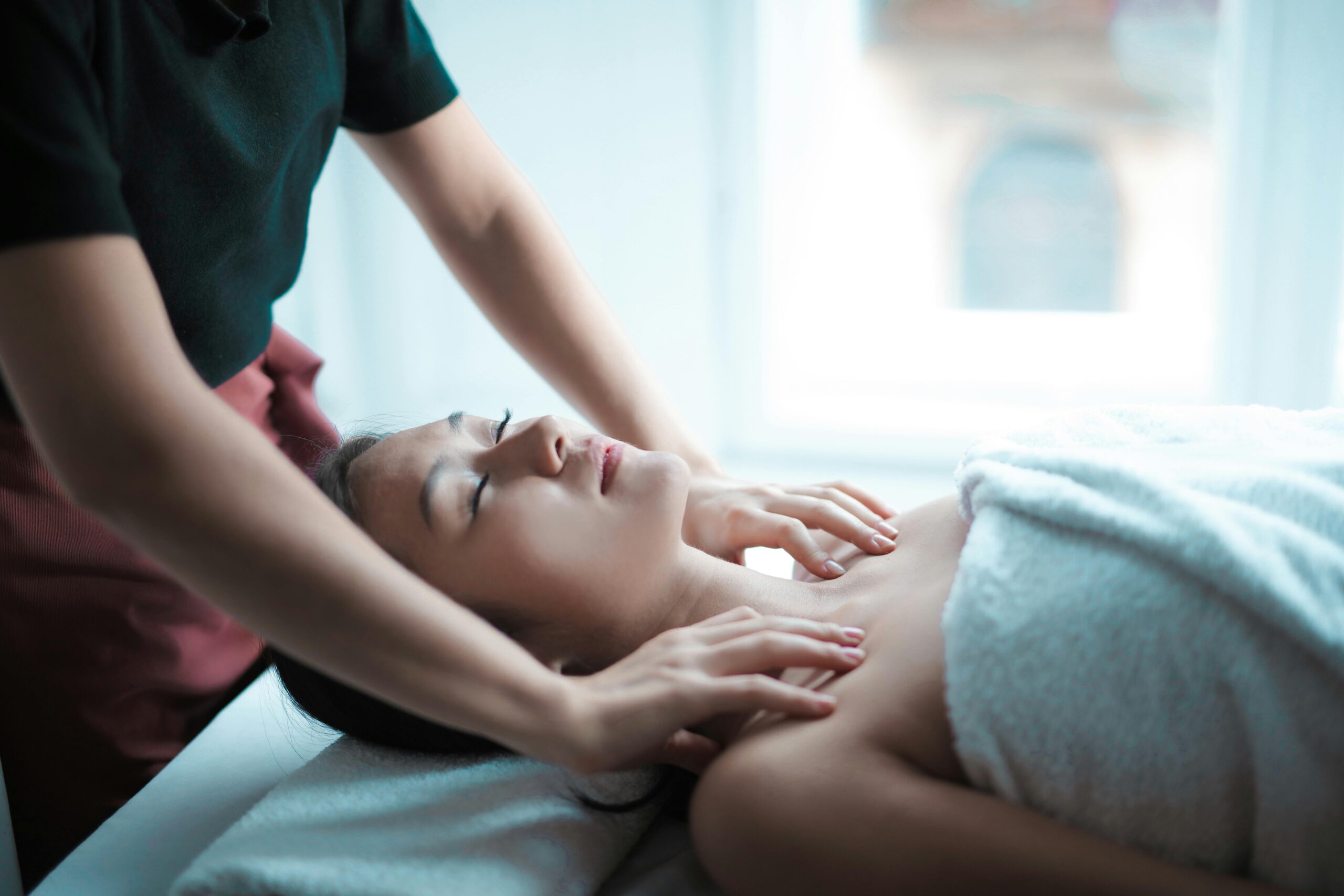 Therapist gently massages a towel‑wrapped woman’s neck and shoulders as she lies on her back.