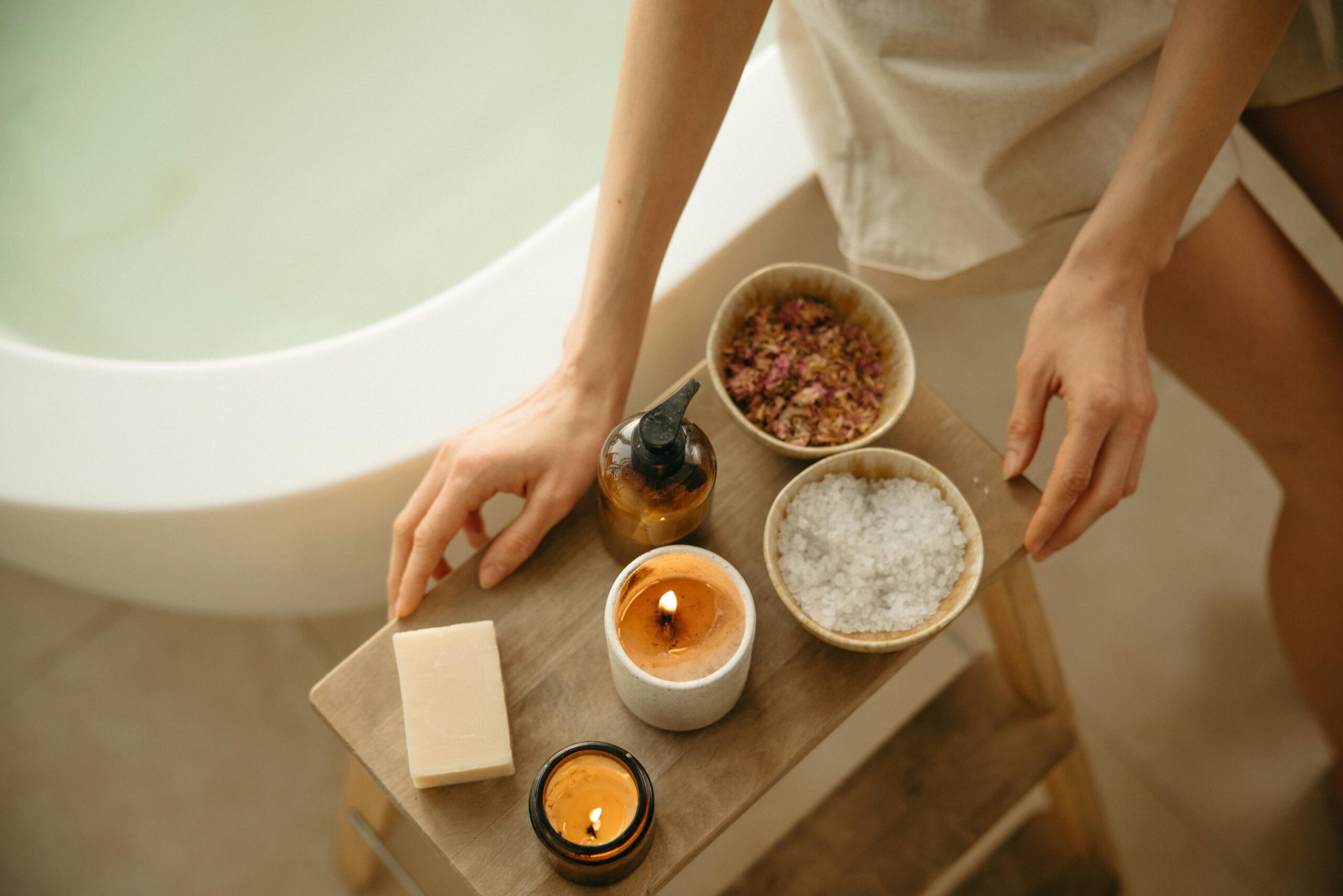 Overhead view of spa essentials—soap, candles, bath salts, and dried petals—arranged on a wooden stool by a tub.