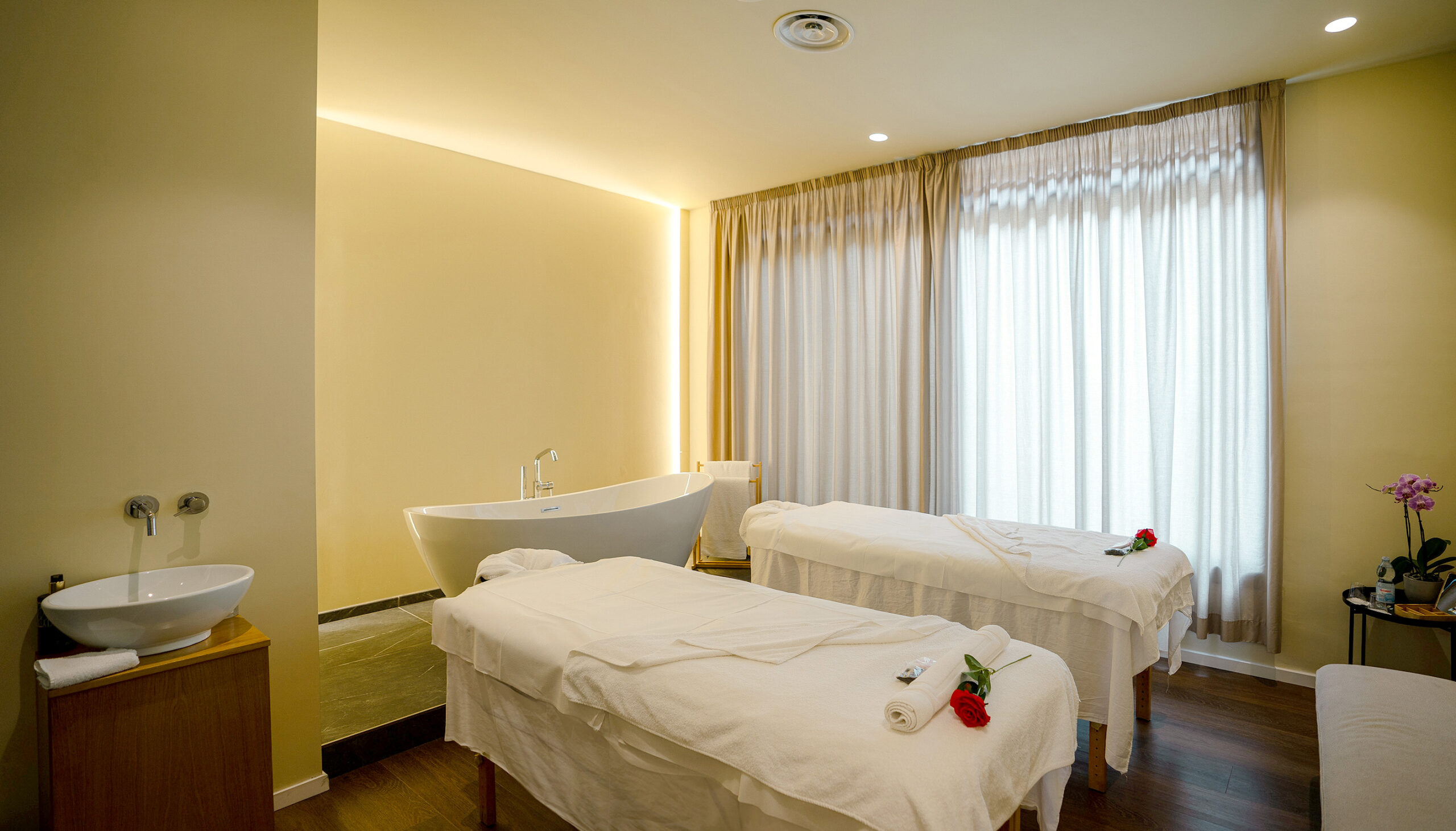Peaceful treatment room featuring two massage tables beside a freestanding soaking tub and sheer curtained window.